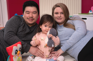 Image shows a mum, dad, and their daughter who has SMA, sitting down on the ground. The child is holding a cuddly toy.