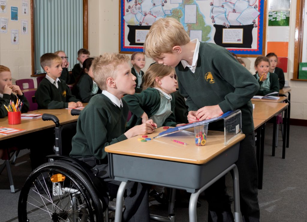boy with SMA in manual chair at desk in classroom chatting with friend