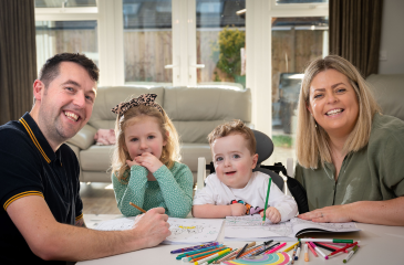 Image shows a family sitting round a table colouring - a mum, dad, daughter and their son who has SMA Type 1.