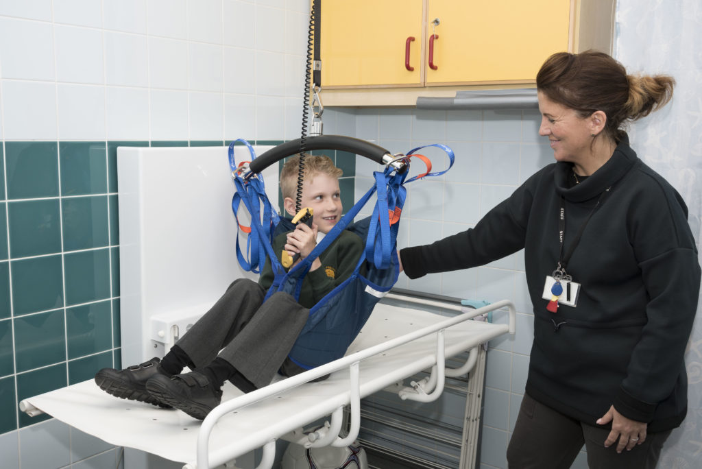Image shows a young boy who has SMA using a hoist with a PA assisting him.