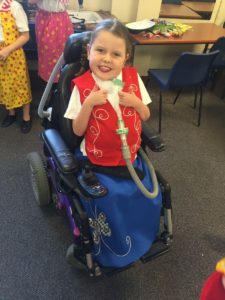 Image shows a young girl who has SMARD sitting in her powerchair. She has a tracheostomy, and is wearing a red top and a blue skirt.