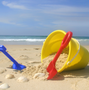 Image shows blue sky and sea and golden sand. There's a red and a blue spade with a yellow bucket.