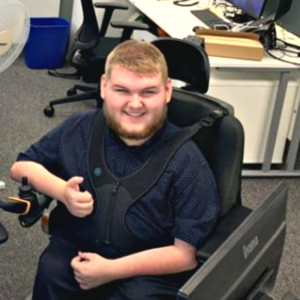 Image shows a young adult, who has SMA, sitting in his wheelchair in his office, giving a thumbs up.