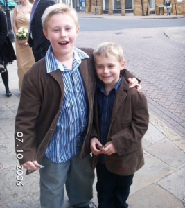 Image shows two young boys standing with their arms around each other. They are both wearing brown jackets, jeans and a blue shirt.