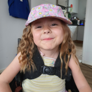 Image shows a young girl who has SMA, sitting in her wheelchair. She has brown hair, and is wearing a yellow top and a pink hat.