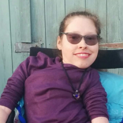 Image shows an adult woman, who has SMA, sitting in her wheelchair and wearing a dark purple top.