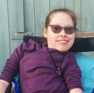 Image shows an adult woman, who has SMA, sitting in her wheelchair and wearing a dark purple top.