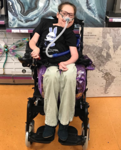 Image shows an adult woman, who has SMA, sitting in her wheelchair. She has a breathing mask on, and is wearing a dark t-shirt and cream trousers.