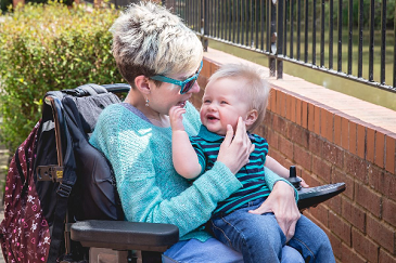Image shows an adult woman who has SMA, sitting in her wheelchair holding her baby boy. Both have blonde hair and are wearing blue.