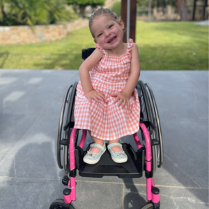 Image shows a little girl who has SMA, sitting in her wheelchair and smiling at the camera. She has blonde hair and is wearing a pink dress.