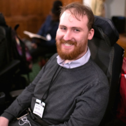 Image shows an adult man, who has SMA, sitting in his wheelchair and smiling at the camera. He's wearing a dark grey jumper.