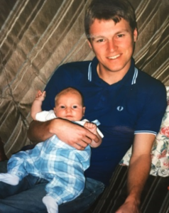 Image shows an adult man wearing a dark blue t-shirt. He is holding his baby boy who is in a light blue baby grow.