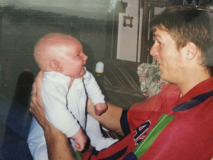 Image shows a man in a red t-shirt holding his baby boy and smiling.