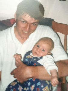 Image shows a man in a white t-shirt holding his baby boy and smiling at the camera.