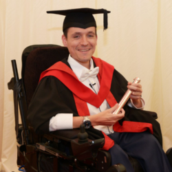 Image shows an adult man, sitting in his powerchair, at his graduation. He's wearing black and red robes, and a mortarboard.