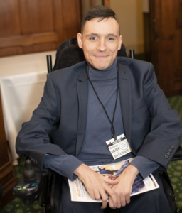 Image shows an adult man who has SMA, sitting in his wheelchair. He's wearing a dark polo neck jumper and dark jacket.