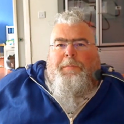 Image shows an adult man, who has SMA, sitting in his wheelchair. He has grey hair and a grey beard, and is wearing a blue hoodie.