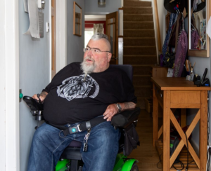 Image shows an adult man who has SMA driving around his hallway in his wheelchair.