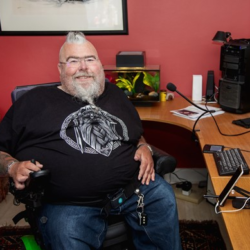Image shows an adult man who has SMA, sitting in his wheelchair at his desk, surrounded by assistive technology.