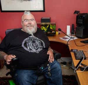 Image shows an adult man who has SMA, sitting in his wheelchair at his desk, surrounded by assistive technology.