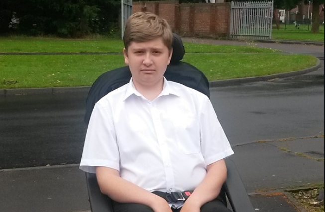 Image shows a teenager who has SMA, sitting in his wheelchair. He is wearing a white shirt and grey trousers.