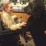 Image shows an adult woman, who has SMA, sitting in her wheelchair. She is holding the paw of her black goldendoodle dog.