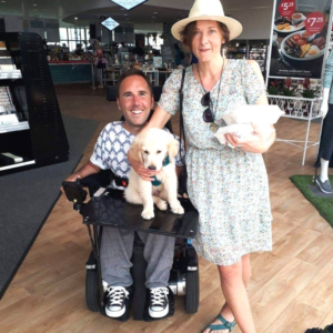 Image shows an adult man, who has SMA, with a golden retriever puppy sitting on his lap in his wheelchair. They are next to his partner who is standing and wearing a green dress.