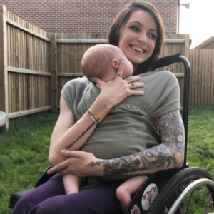 Image shows an adult woman who has SMA, sitting in her wheelchair. She is holding her baby boy.