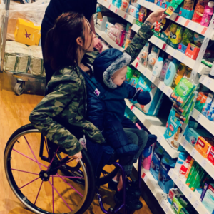 Image shows an adult woman at the shops. She is in her wheelchair with her baby son on her lap, as they browse the shelves.