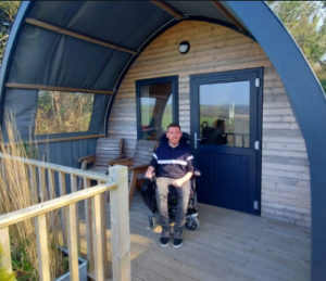 Image shows an adult man witting in his powerchair outside an accessible cabin.