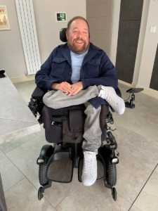 Image shows an adult man sitting in his wheelchair, with one leg crossed over the other. He is wearing grey trousers and a blue shirt.