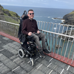 Image shows an adult man, who has SMA, sitting in his wheelchair with a view behind him of the sea.