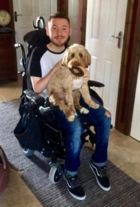 Image shows an adult man who has SMA, sitting in his wheelchair. His golden dog is sitting on his lap.