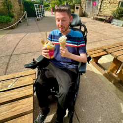 Image shows an adult man, who has SMA, sitting in his wheelchair holding an ice cream in one hand, and a drink in the other.