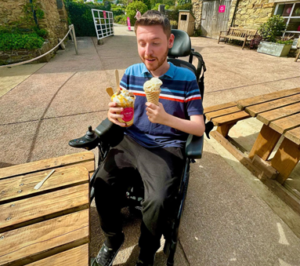 Image shows an adult man, who has SMA, sitting in his wheelchair holding an ice cream in one hand, and a drink in the other.