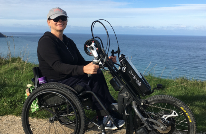 Images shows an adult woman using her handcycle in front of a sea view.