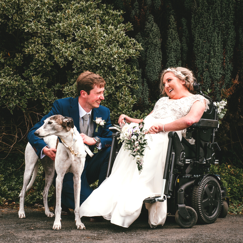Image shows a lady who has SMA sitting in her wheelchair wearing a wedding dress and holding a bunch of flowers. She is next to a man who is crouching down, patting a greyhound.