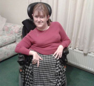 Image shows an adult lady, who has SMA, sitting in her wheelchair. She is wearing a pink jumper and a black and white skirt.