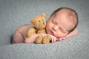 Image shows a baby boy lying down, holding a teddy bear.