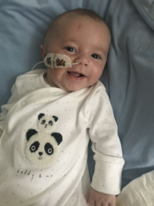 Image shows a baby boy lying down. He is wearing a white baby grow with a panda picture on it.