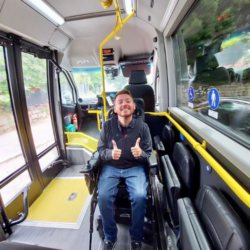 Image shows an adult man, who has SMA, sitting in his wheelchair on a bus, giving a thumbs up.