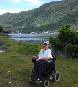 Image shows a lady who has SMA sitting in her wheelchair. She is on holiday, in front of a large lake and mountains. She is wearing a cream baseball cap, a white t-shirt and black trousers.