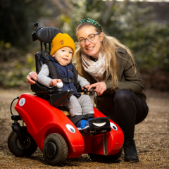 Image shows a little boy who has SMA Type 1 sitting in his Wizzybug wheelchair. He is next to his mum who is crouching down with her arm round him.
