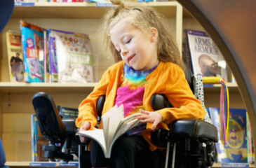 Image shows a young girl with blond hair. She is sitting in her wheelchair and reading a book.