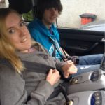 Image shows Suzanne behind the wheel of an adapted car. Her brother is sitting in the passenger seat. They are both smiling at the camera.