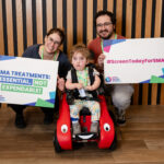 Image shows a couple with a child in a whizzy bug holdig signs that say #ScreenTodayForSMA and SMA treatements: essential not exendable