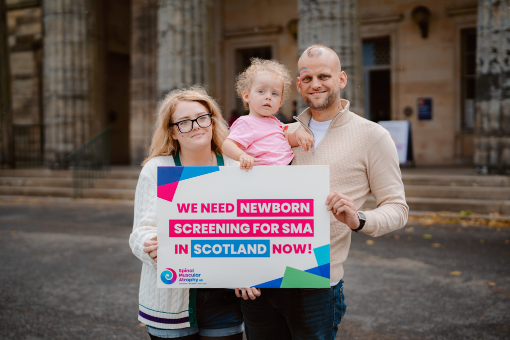 Mum and Dad with holding their young daughter with an SMA UK placard