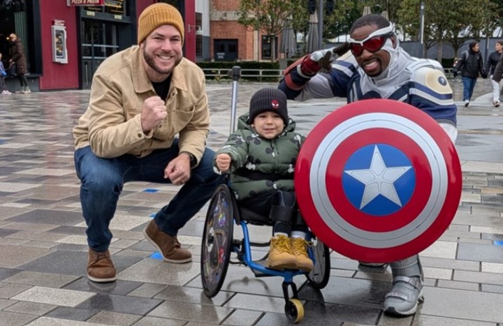Boy in a wheelchair with his father on one side and Captain America holding a shield on the other